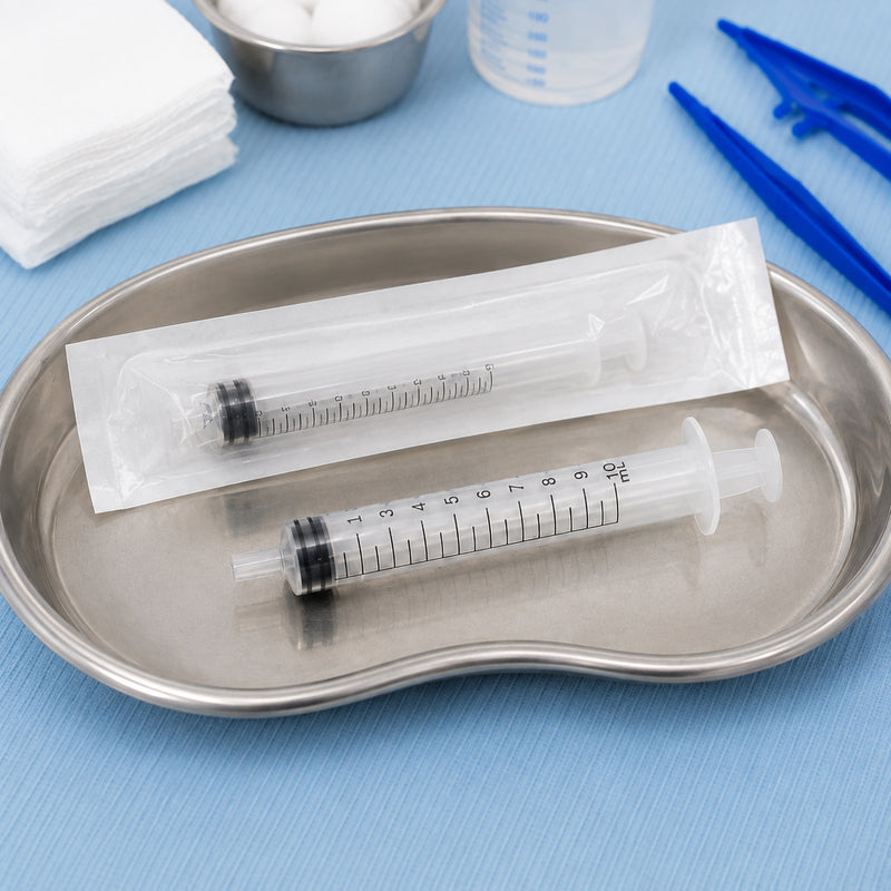 Two AeroSupplies syringes presented in a stainless steel kidney dish on a prepared clinical surface. One syringe remains inside its individual sterile wrapper while the second is unpackaged, clearly showing the transparent barrel, black graduated scale, plunger and luer slip connection.