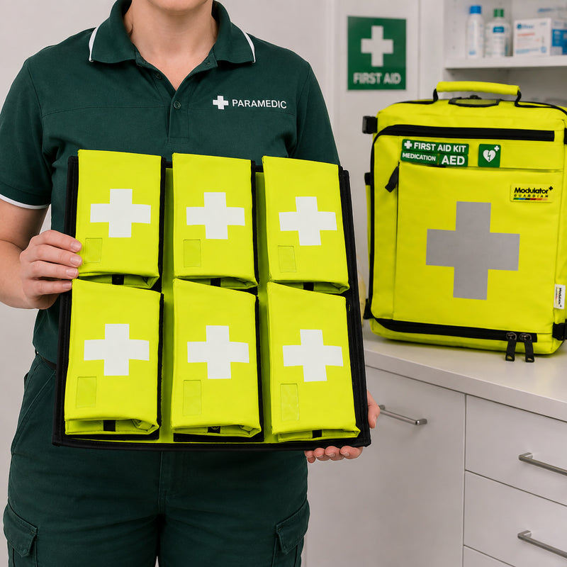 Paramedic holding the Aero Healthcare Allergy Board Insert with 6 high visibility first aid kit bags arranged in two rows of three. An AeroKit Modulator Guardian first aid backpack is visible in the clinical background, demonstrating how the removable board can support organised allergy and emergency supply storage.