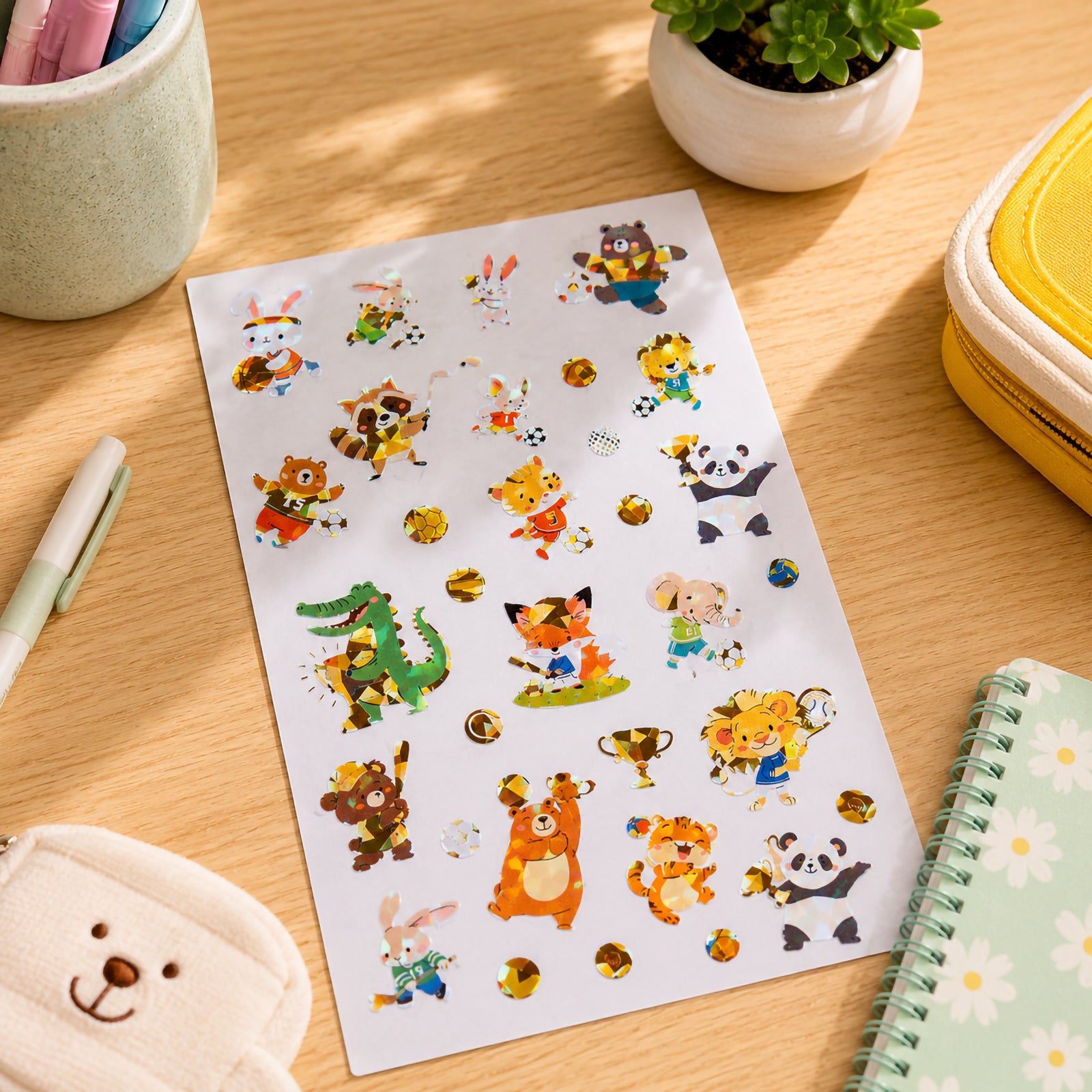Full sticker sheet photographed on a desk surrounded by stationery and classroom supplies. The colourful sports themed animals and shiny foil balls, trophies and equipment create an engaging activity for children.