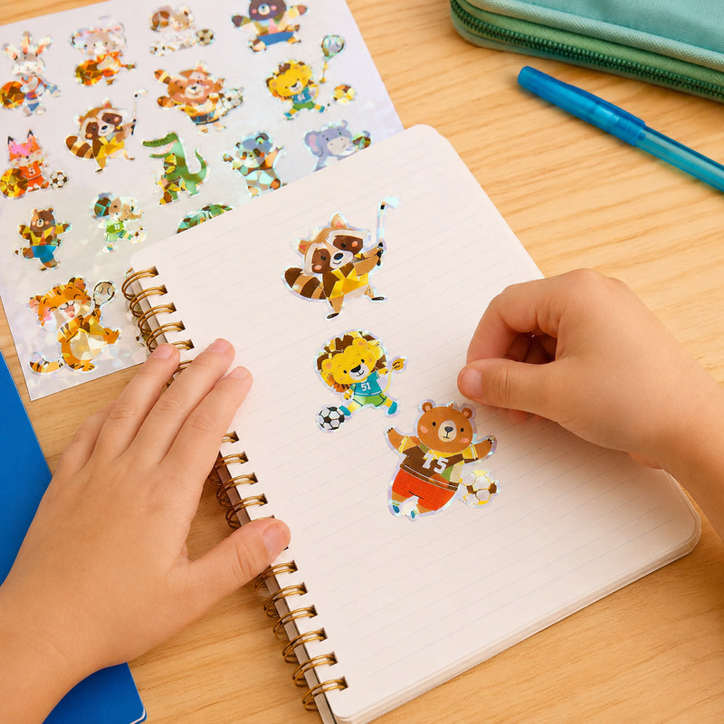 Child decorating a notebook with holographic animal athlete stickers featuring a raccoon hockey player, lion soccer player and bear football character. The sparkling foil designs encourage creative play and personalisation.