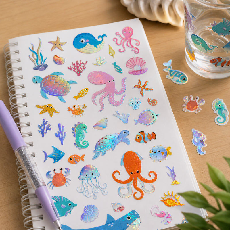 Full sheet of Aquatic Adventures holographic stickers displayed on a notebook beside stationery and decorative items. The collection includes whales, turtles, octopuses, seahorses, fish, coral and shells with sparkling foil details.
