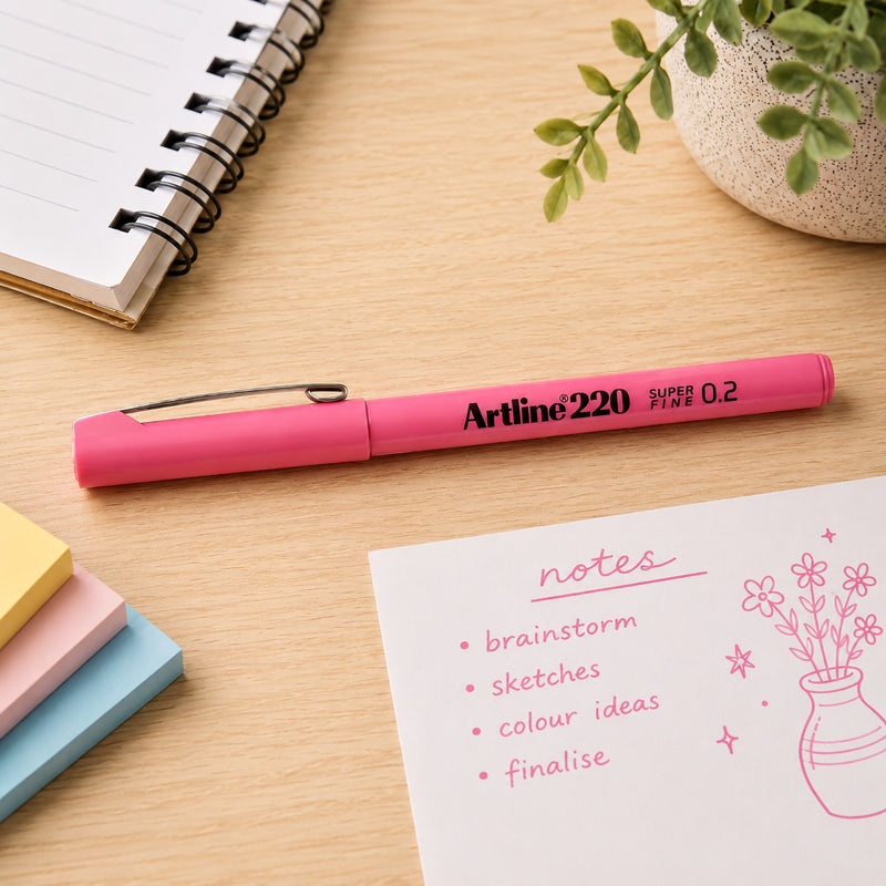 The Artline 220 Fineliner Pen 0.2mm Pink rests on a modern wooden desktop beside pastel sticky notes, a notebook and handwritten pink notes. The capped pen is displayed prominently to showcase its slim barrel, metal clip and vibrant pink finish in an organised office environment.