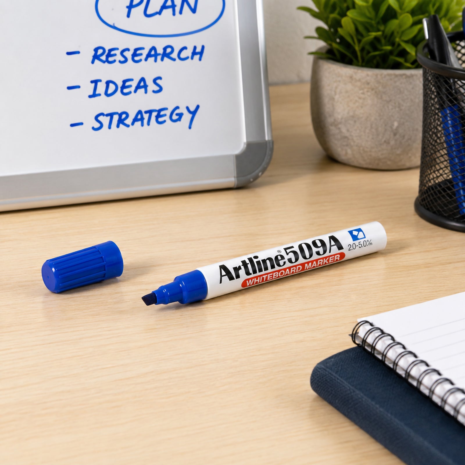 Lifestyle desktop scene featuring the Artline 509A Blue Whiteboard Marker positioned in front of a whiteboard with planning notes. The organised workspace with notebook, pen holder and plant demonstrates the marker's suitability for office planning, education and everyday productivity.