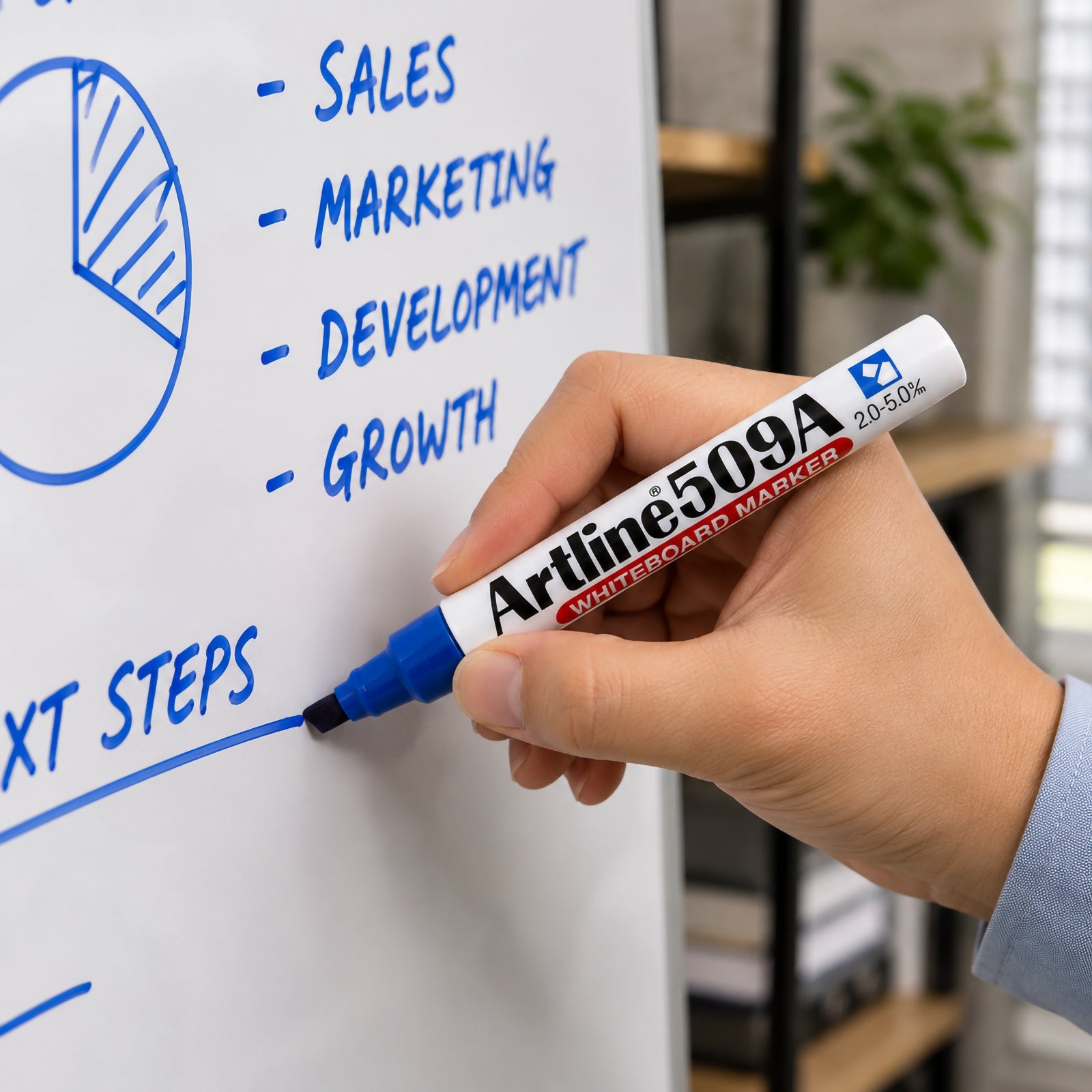 Close up lifestyle image of an Artline 509A Blue Whiteboard Marker being used to write clear blue text on a whiteboard during a business planning session. The chisel nib creates bold, legible writing while the modern office background reinforces its professional use for meetings, presentations and collaborative work.