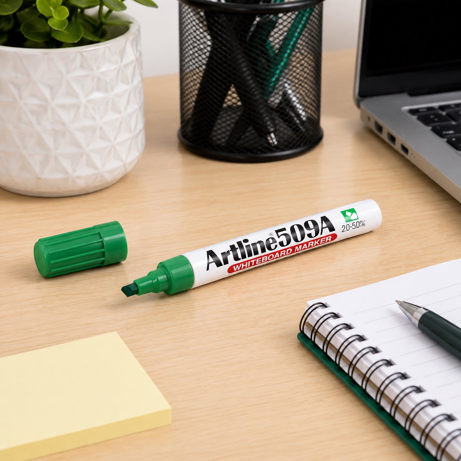 Desktop lifestyle image featuring the Artline 509A Green Whiteboard Marker alongside a notebook, laptop, stationery holder and decorative plant. The clean office environment demonstrates the marker as a reliable choice for meetings, study and everyday business use.