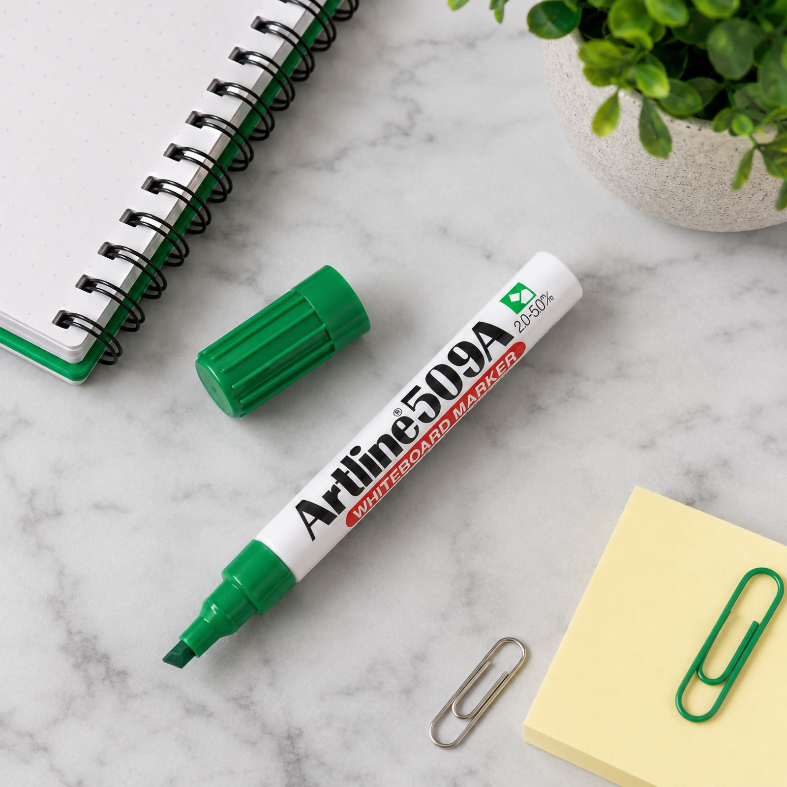 Top view of the Artline 509A Green Whiteboard Marker resting beside an open notebook, sticky notes, paper clips and a potted plant on a marble desktop. The organised workspace highlights the marker as a practical tool for planning, note taking and office productivity.
