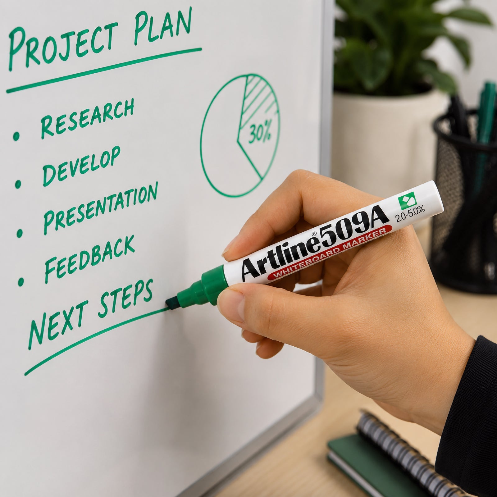 Lifestyle close up showing the Artline 509A Green Whiteboard Marker writing a project plan on a whiteboard. The bold green writing and chisel nib demonstrate clear visibility, making it ideal for presentations, planning sessions and classroom collaboration.