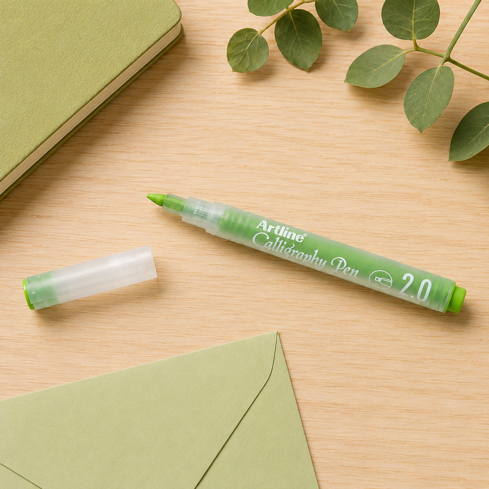 Top view of the Artline Calligraphy Pen 2.0mm Pastel Lime Green on a timber desktop with matching stationery and envelope. The pen is shown uncapped beside its translucent cap, emphasising the broad calligraphy tip and soft pastel colour for creative writing applications.