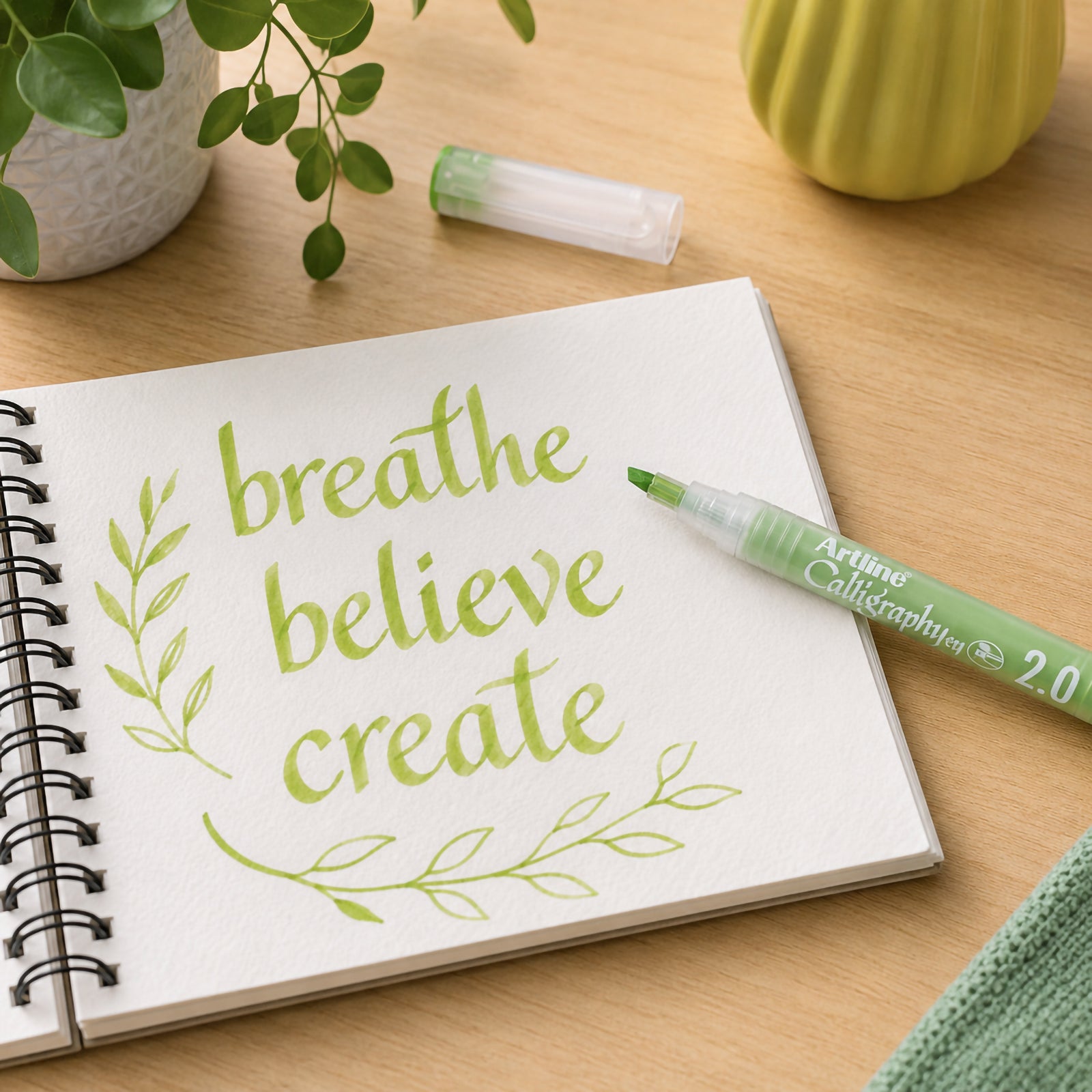 The Artline Calligraphy Pen 2.0mm Pastel Lime Green rests beside an open sketchbook featuring decorative calligraphy and botanical flourishes. The uncapped pen displays its broad nib while the calming workspace demonstrates its suitability for lettering, creative projects and artistic expression.