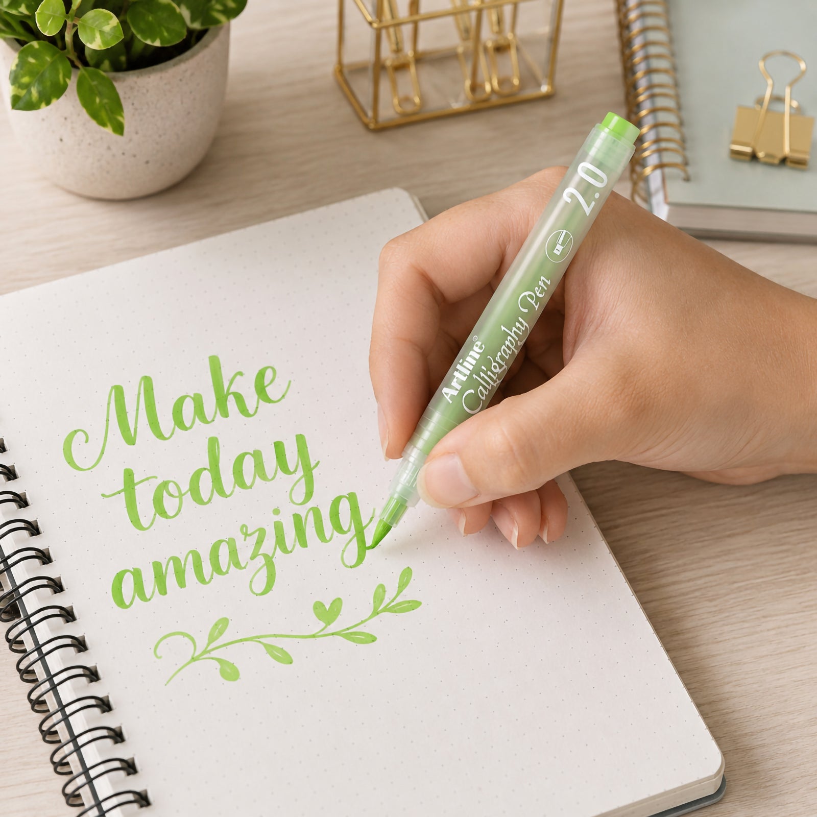 A hand uses the Artline Calligraphy Pen 2.0mm in Pastel Lime Green to create elegant hand lettering inside a dotted notebook. The soft pastel ink produces smooth, bold strokes, surrounded by a modern desktop with stationery and indoor plants that highlights creative journalling and calligraphy.