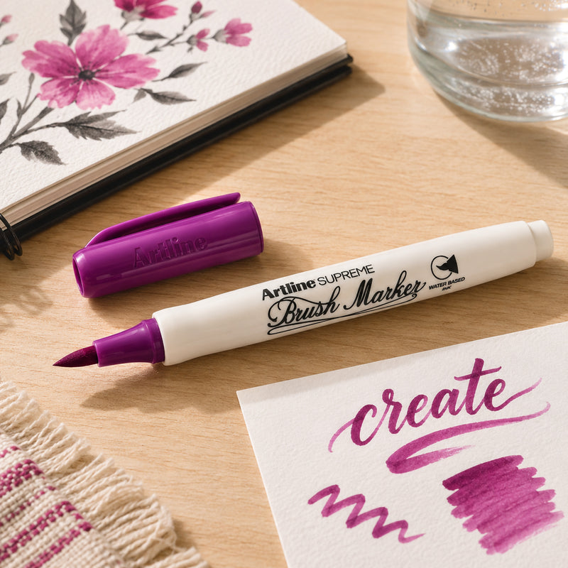The Artline Supreme Brush Marker Magenta is displayed uncapped on a wooden desk beside a sketchbook, floral artwork and lettering samples. The image highlights the vibrant magenta ink, flexible brush tip and comfortable ergonomic barrel designed for creative expression.