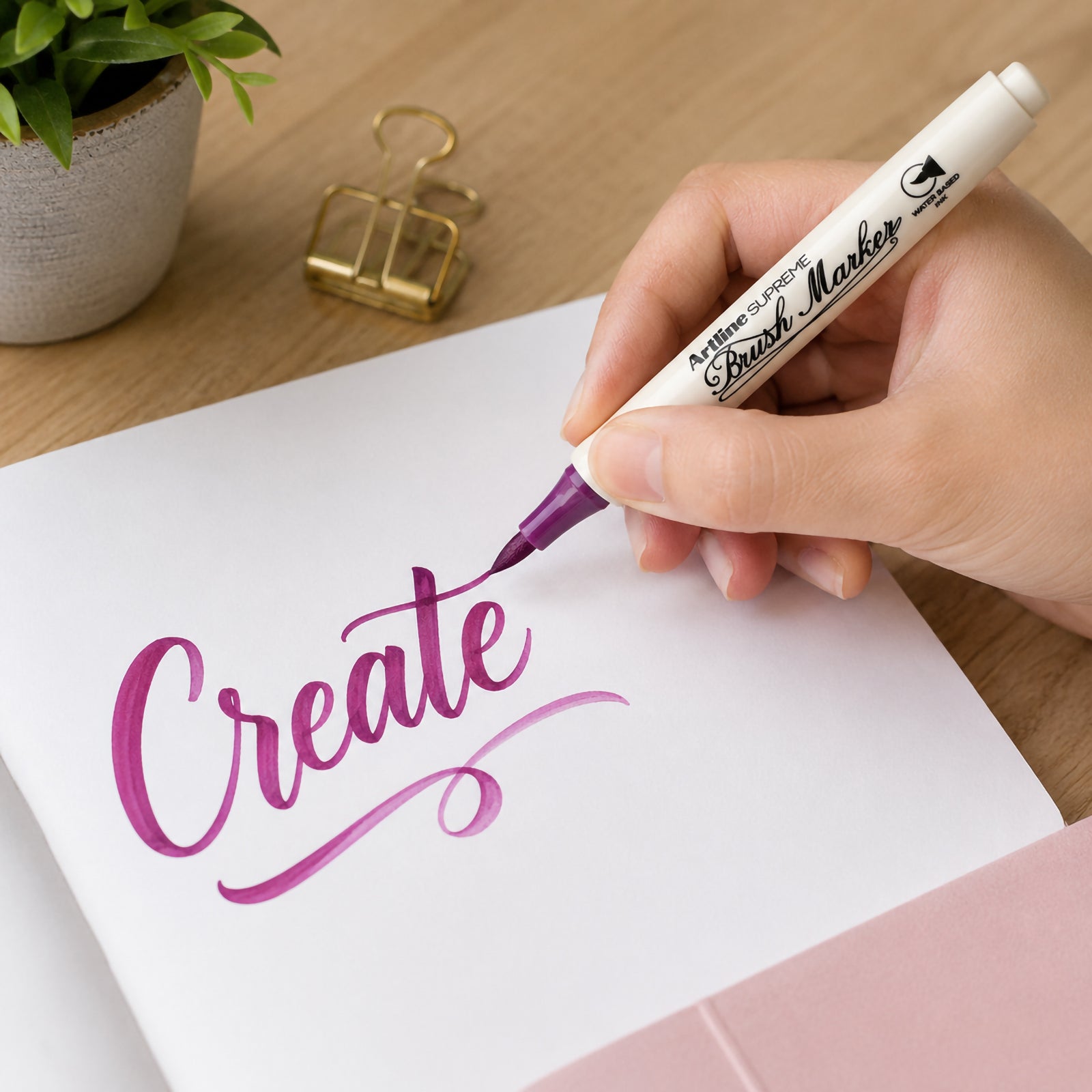 A person uses the Artline Supreme Brush Marker Magenta to create elegant brush lettering on white paper. The flexible brush tip produces smooth thick and thin strokes, demonstrating precise control for modern calligraphy, creative journalling and artistic lettering.