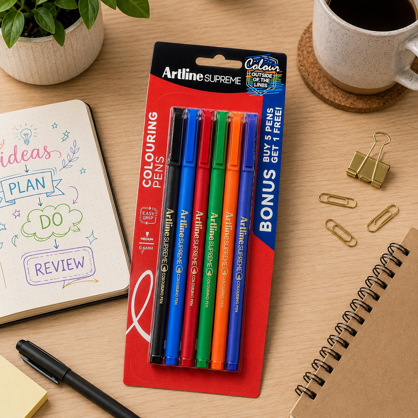 The Artline Supreme Colouring Pen assorted pack is displayed on a tidy workspace beside notebooks, stationery and coffee. The retail packaging showcases the six vibrant pens and promotes the bonus pack presentation for creative users.