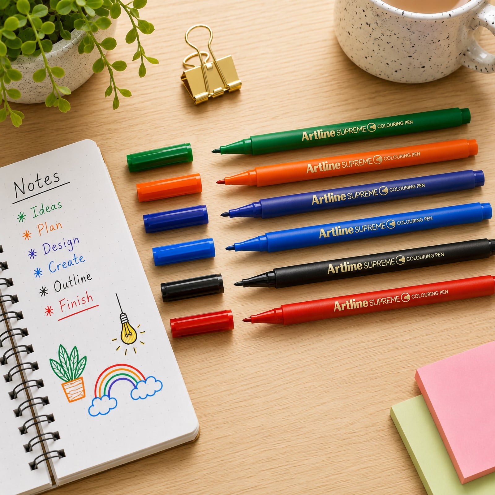 Six Artline Supreme Colouring Pens in assorted colours are arranged neatly on a wooden desk beside an open notebook filled with colourful notes and simple illustrations. The image highlights the vibrant colour selection and fine fibre tips.