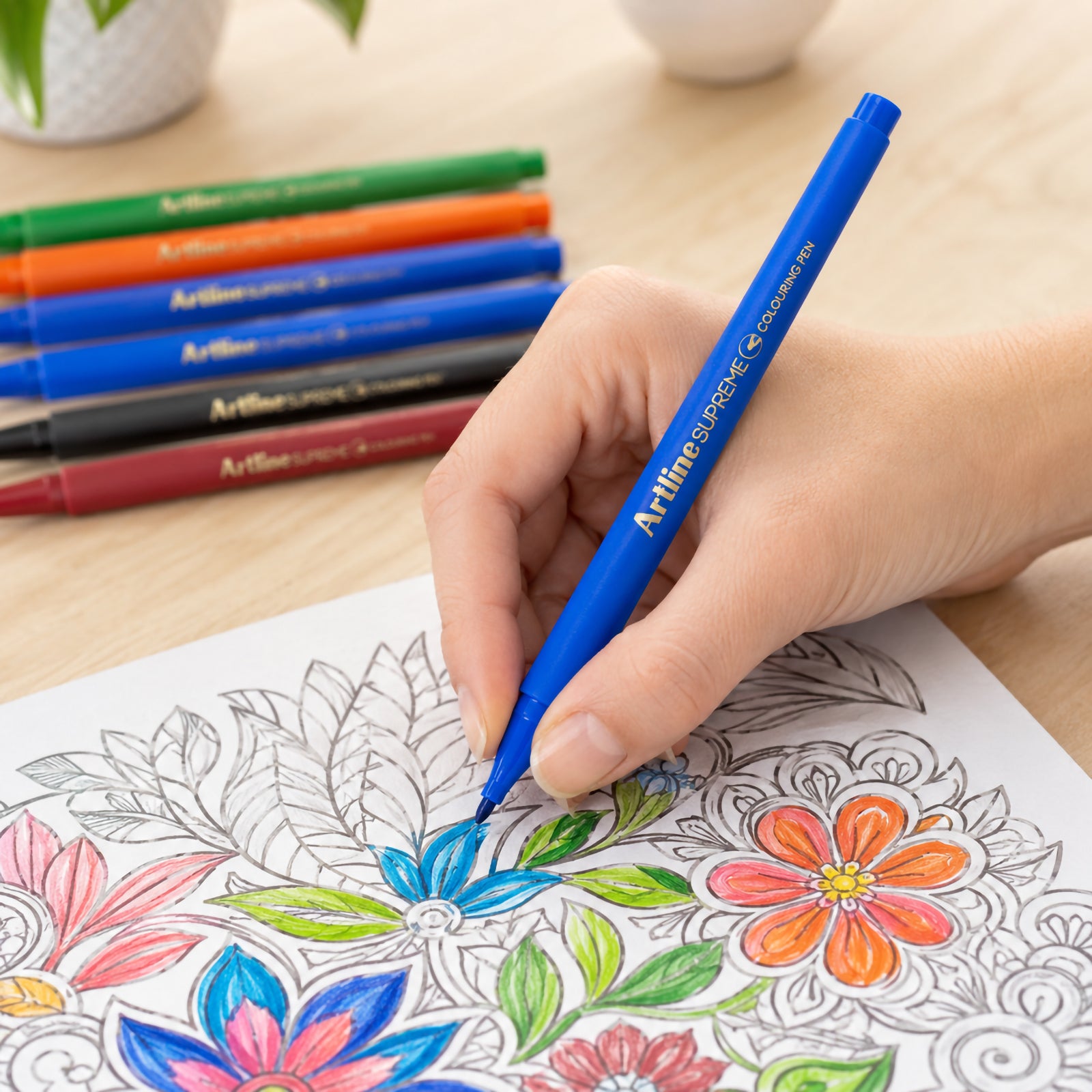 A hand uses the blue Artline Supreme Colouring Pen to colour intricate floral artwork on a colouring page. The close up lifestyle image demonstrates the fine 0.6mm fibre tip delivering precise colour application for detailed creative projects.