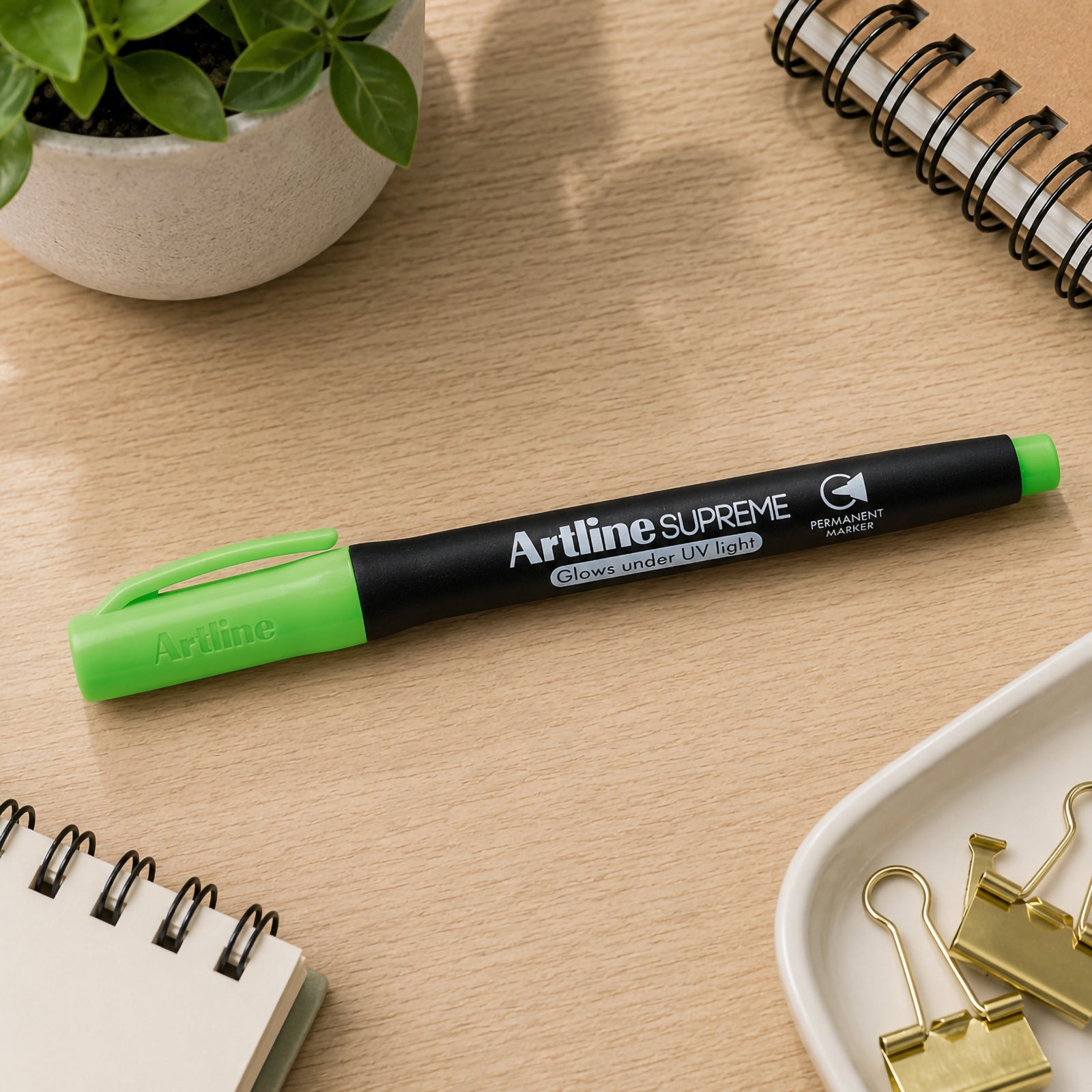 The capped Artline Supreme Glow Permanent Marker Green rests across a wooden desk beside stationery, notebooks and binder clips. The lifestyle setting showcases the marker as an attractive everyday writing tool for creative work, organisation and office use.