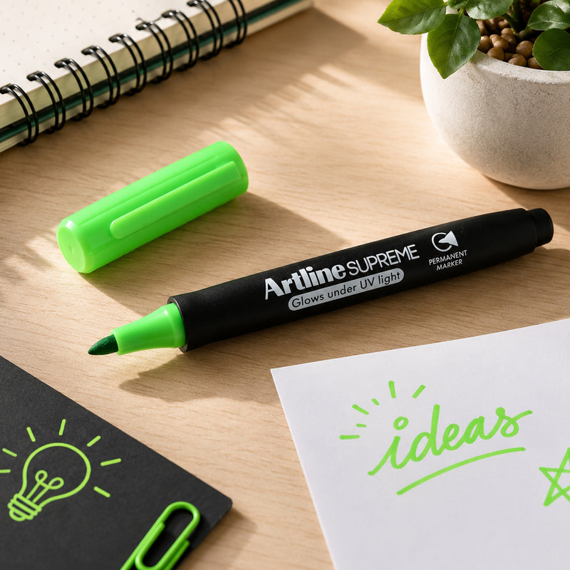 The Artline Supreme Glow Permanent Marker Green is displayed uncapped beside its bright green cap on a tidy desktop with notebooks, paper, decorative plants and office accessories. The image highlights the 1mm bullet tip, premium barrel design and unique UV glow feature.