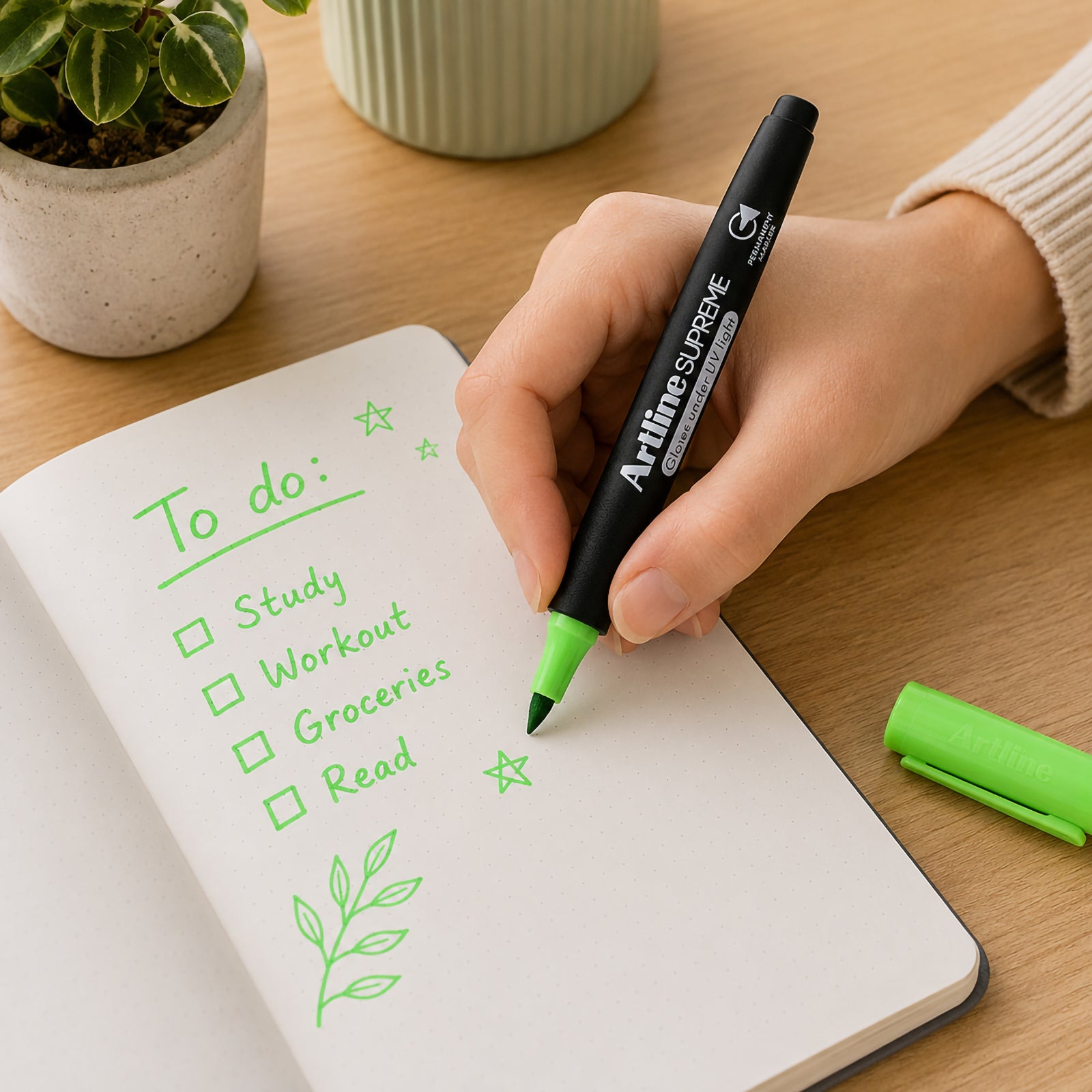 A person uses the Artline Supreme Glow Permanent Marker Green to write a colourful to do list and decorative illustrations inside a notebook. The vivid green permanent ink stands out clearly while demonstrating the marker's comfortable ergonomic grip and smooth writing performance in a modern workspace.