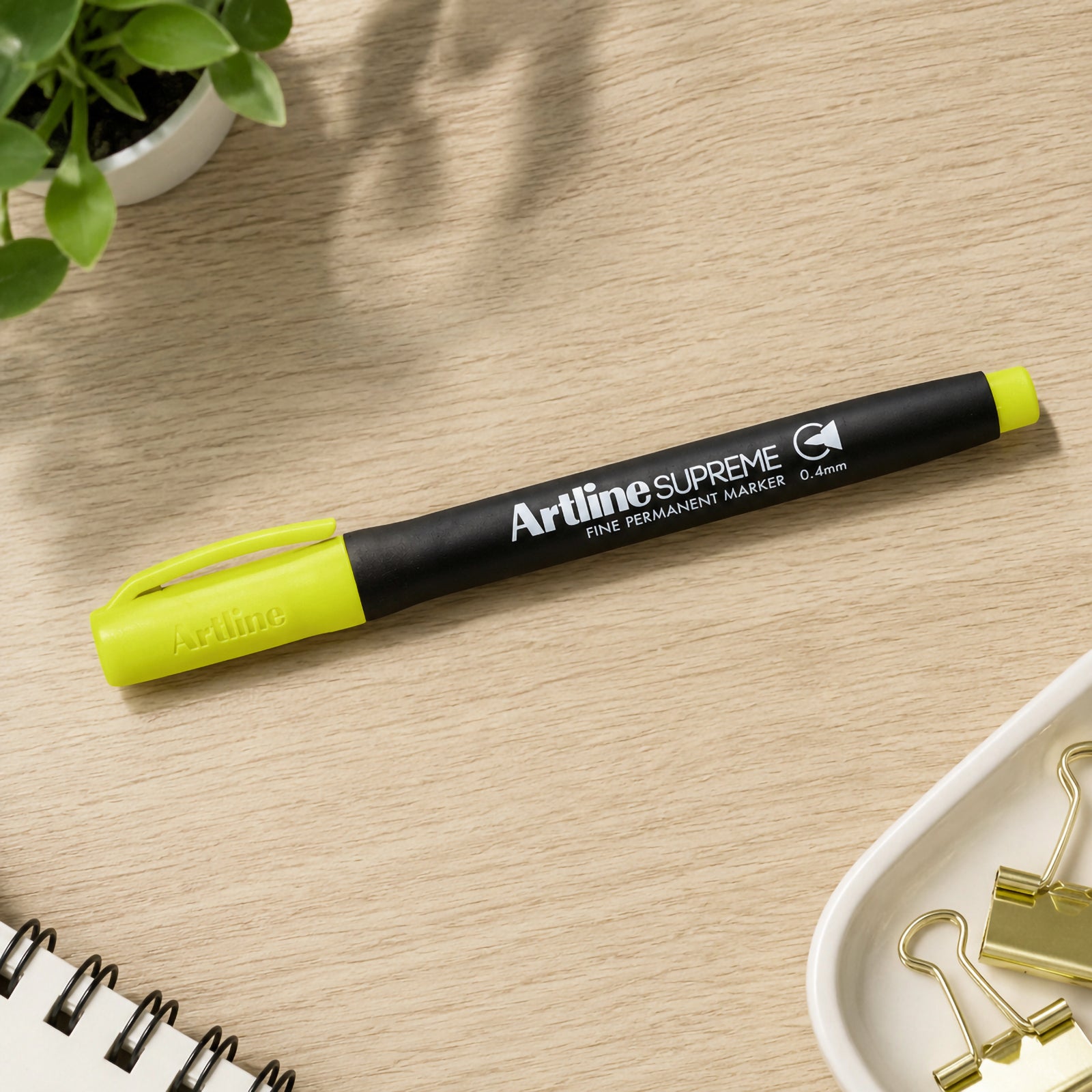The capped Artline Supreme Permanent Marker 0.4mm Blue Yellow rests neatly on a wooden desktop surrounded by stationery, paper clips and a small plant. The lifestyle setting showcases the marker as a colourful addition to organised workspaces and creative planning.