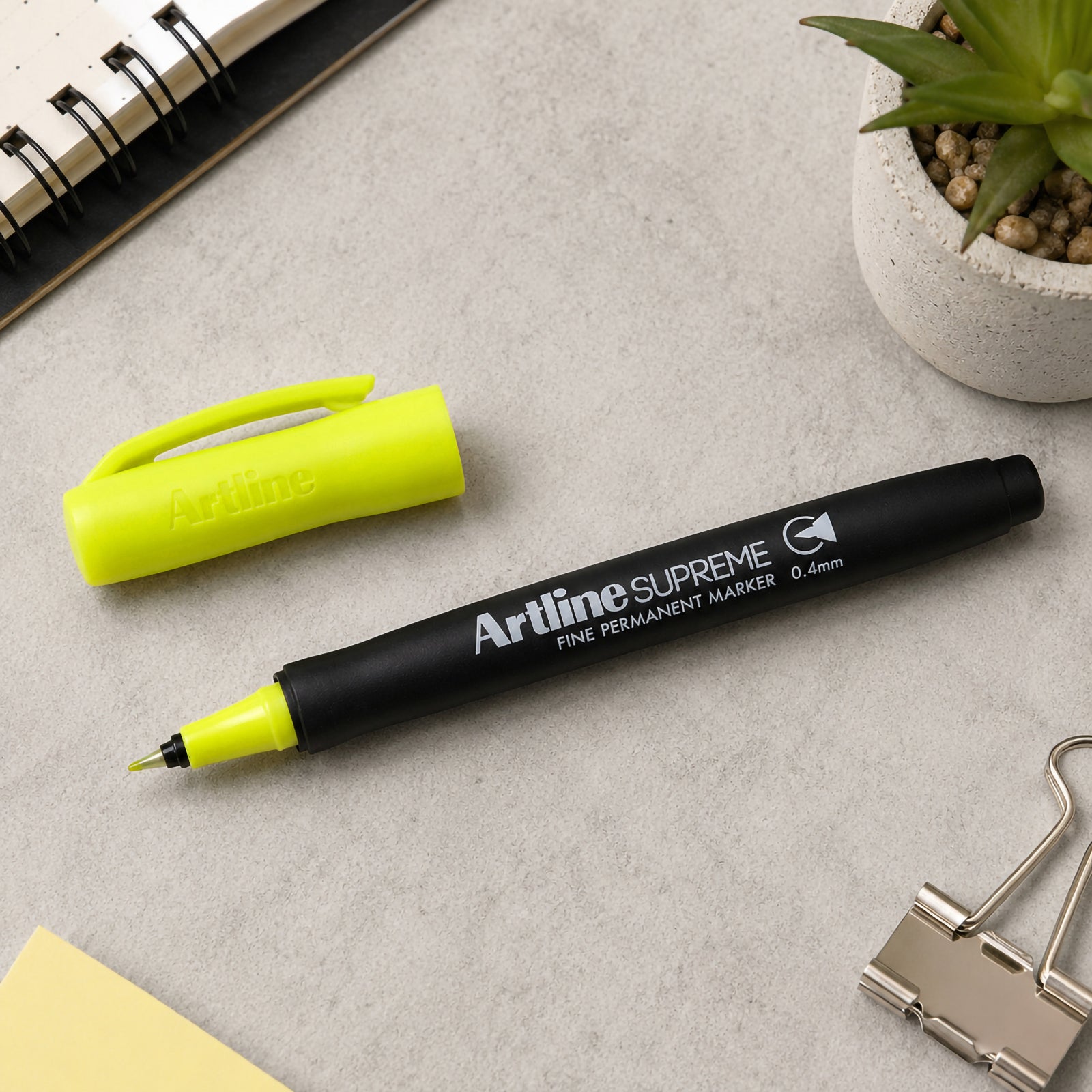 The Artline Supreme Permanent Marker 0.4mm Blue Yellow is displayed uncapped beside its matching fluorescent cap on a clean desktop with a notebook, binder clip and decorative succulent. The image highlights the fine nib, ergonomic barrel and vivid ink colour for detailed marking tasks.