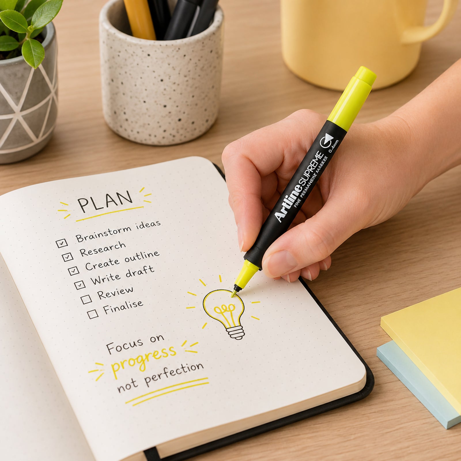 A hand uses the Artline Supreme Permanent Marker 0.4mm Blue Yellow to add bright highlights and illustrations to a notebook planner on a modern office desk. The vibrant yellow ink emphasises important notes and creative icons while stationery and desk accessories create an organised, productive workspace.