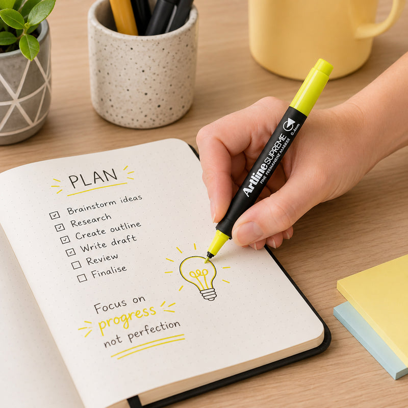 A hand uses the Artline Supreme Permanent Marker 0.4mm Blue Yellow to add bright highlights and illustrations to a notebook planner on a modern office desk. The vibrant yellow ink emphasises important notes and creative icons while stationery and desk accessories create an organised, productive workspace.