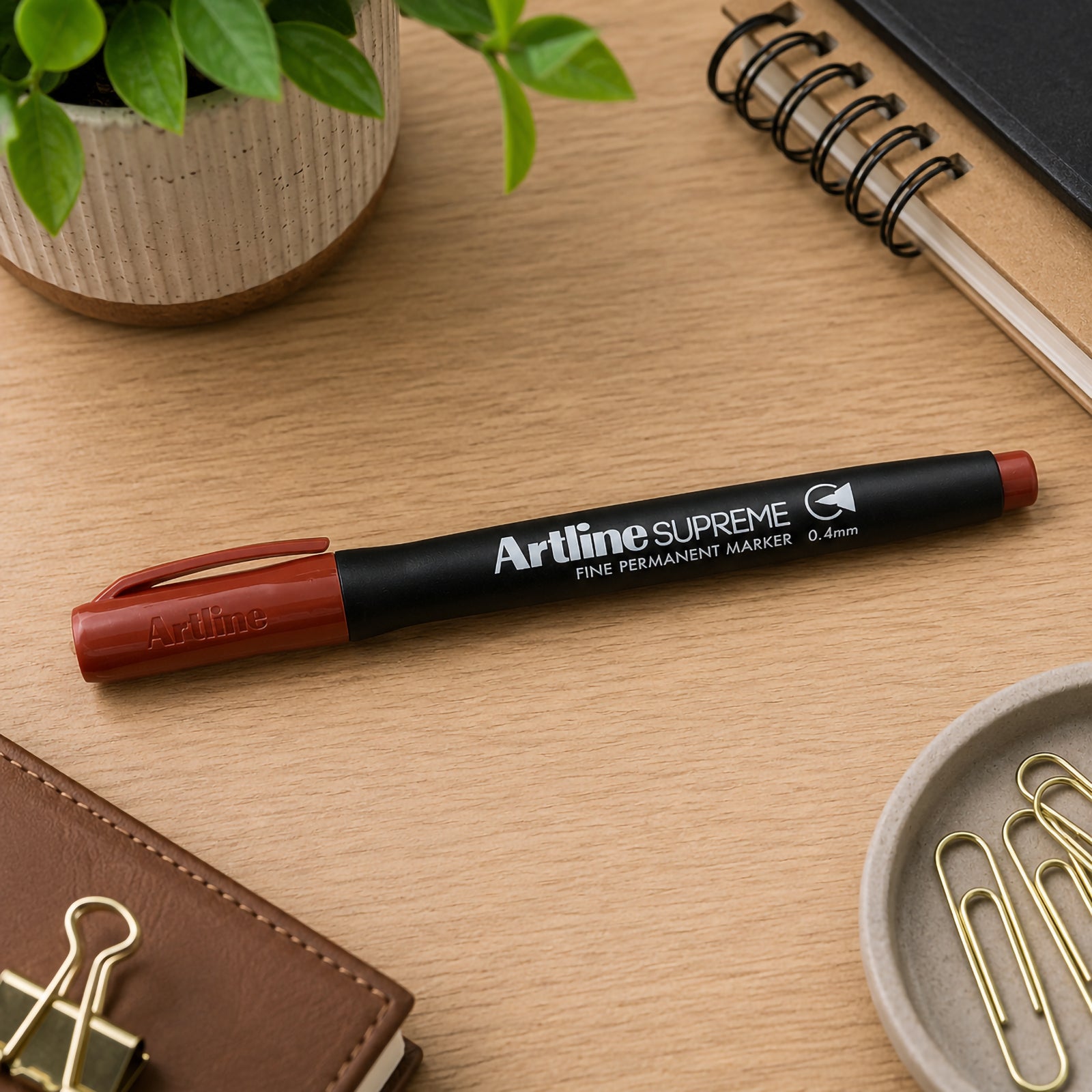 The capped Artline Supreme Permanent Marker 0.4mm Brown is positioned neatly across a tidy office desk surrounded by stationery, notebooks, paper clips and decorative plants. The lifestyle setting showcases the marker as an everyday tool for organised workspaces and creative projects.