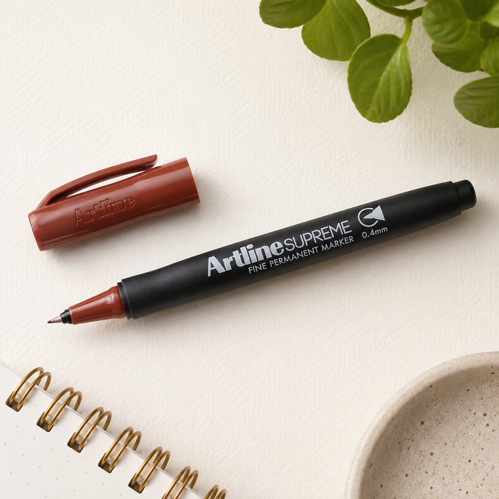 The Artline Supreme Permanent Marker 0.4mm Brown is displayed uncapped beside its matching brown cap on a clean desktop with a notebook, ceramic dish and green foliage. The image clearly highlights the fine writing tip, ergonomic barrel and premium product branding.
