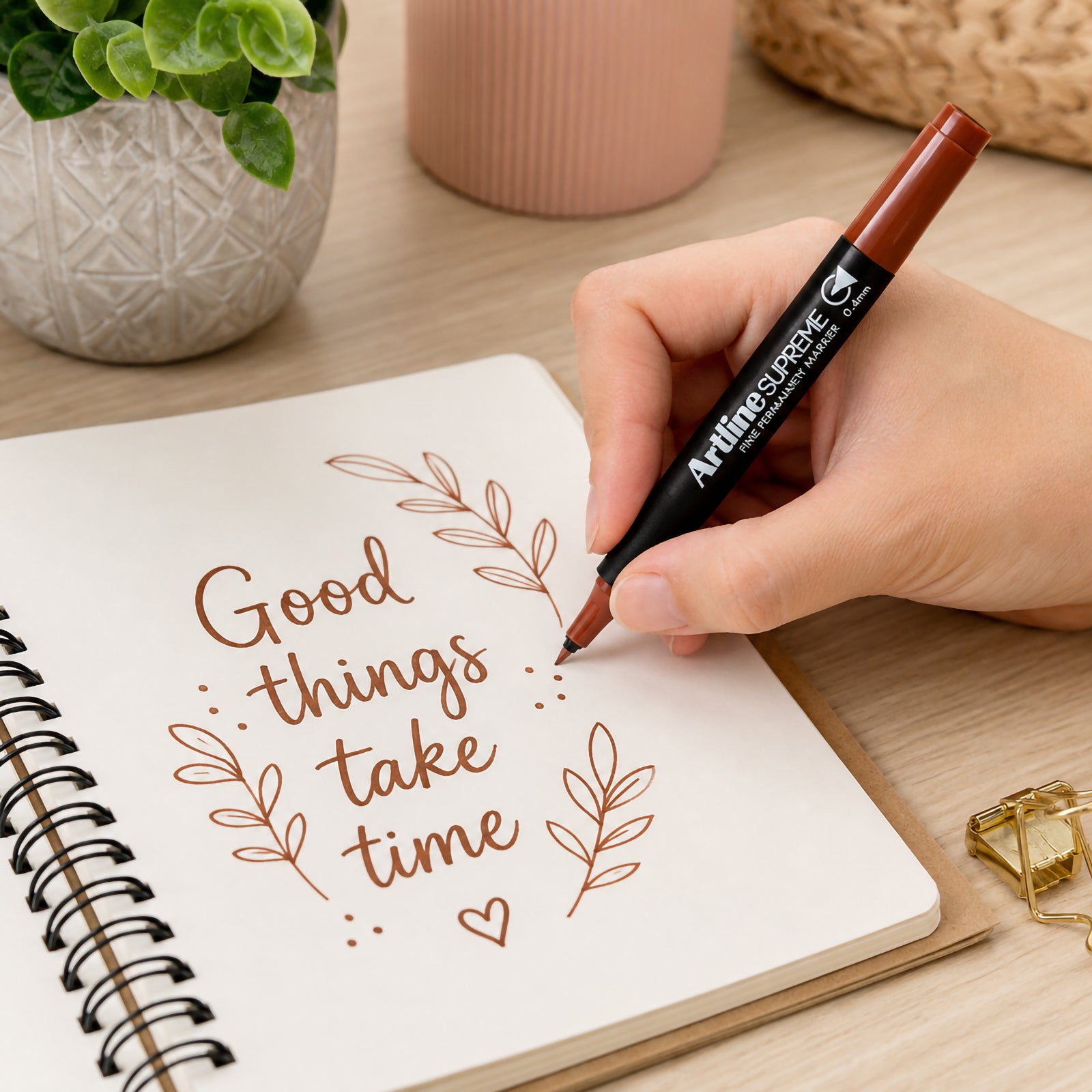 A hand uses the Artline Supreme Permanent Marker 0.4mm Brown to create elegant lettering and decorative leaf illustrations in a spiral notebook on a modern wooden desk. The warm brown ink complements the natural workspace featuring indoor plants and office accessories, demonstrating the marker's precision and smooth writing performance.