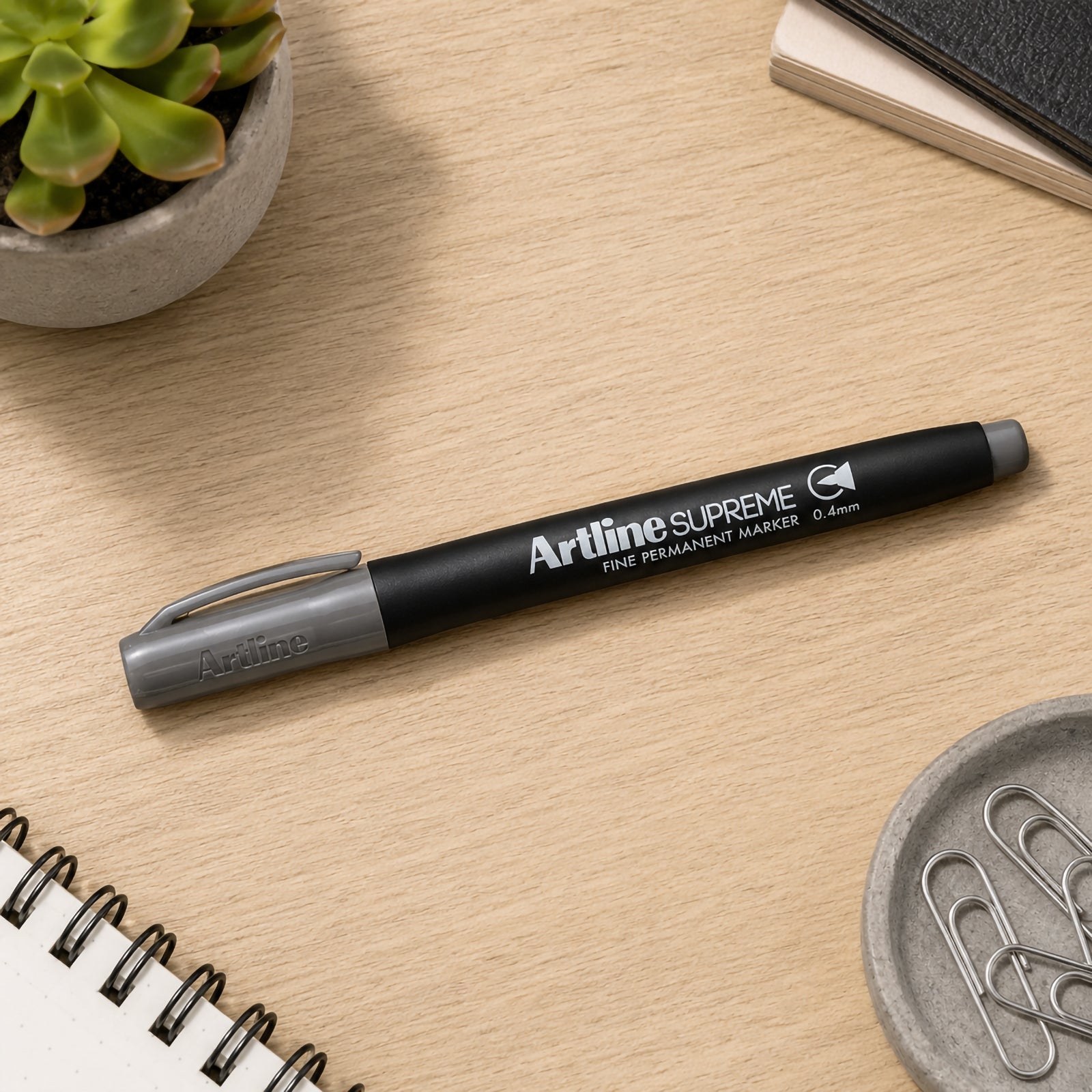 The capped Artline Supreme Permanent Marker 0.4mm Grey rests neatly on a wooden desk beside notebooks, paper clips and a succulent plant. The lifestyle composition presents the marker as an essential everyday writing tool for organised workspaces and creative planning.