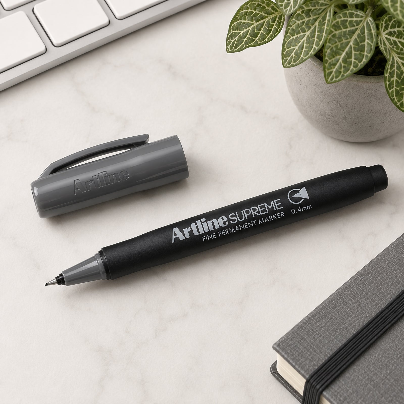 The Artline Supreme Permanent Marker 0.4mm Grey is displayed uncapped beside its matching grey cap on a clean desktop with a notebook, keyboard and decorative plant. The image highlights the fine writing tip, ergonomic barrel and premium branding in a stylish office environment.