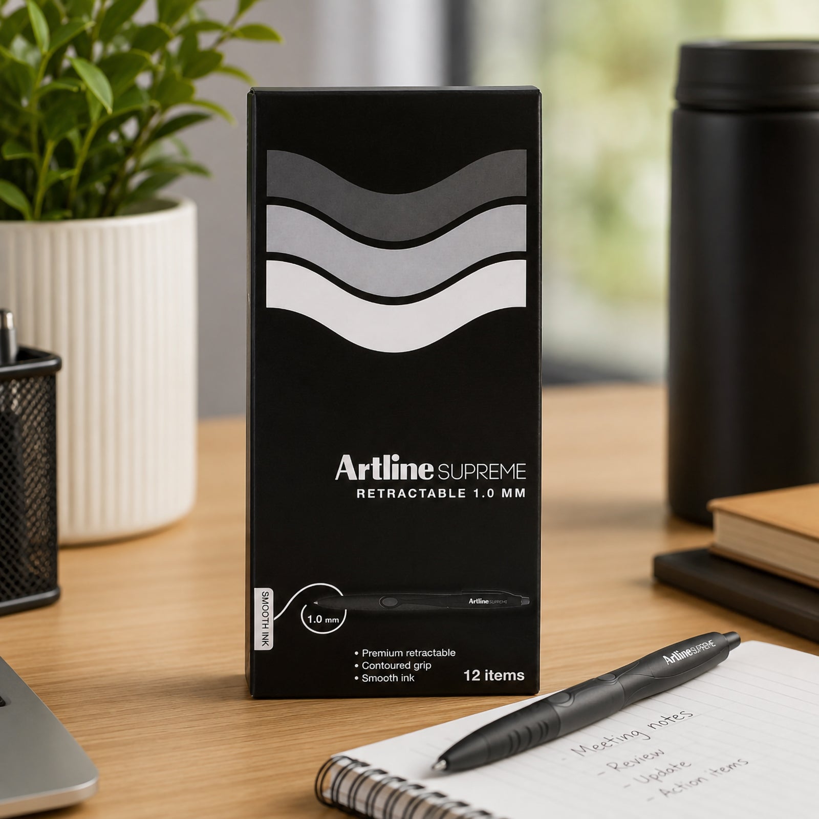 A professional office workspace featuring the Artline Supreme Retractable Ballpoint Pen retail box standing beside an open notebook and matching pen. Surrounded by a laptop, reusable drink bottle and desk accessories, the image presents the premium packaging and demonstrates the product as an ideal choice for business, education and home office environments.