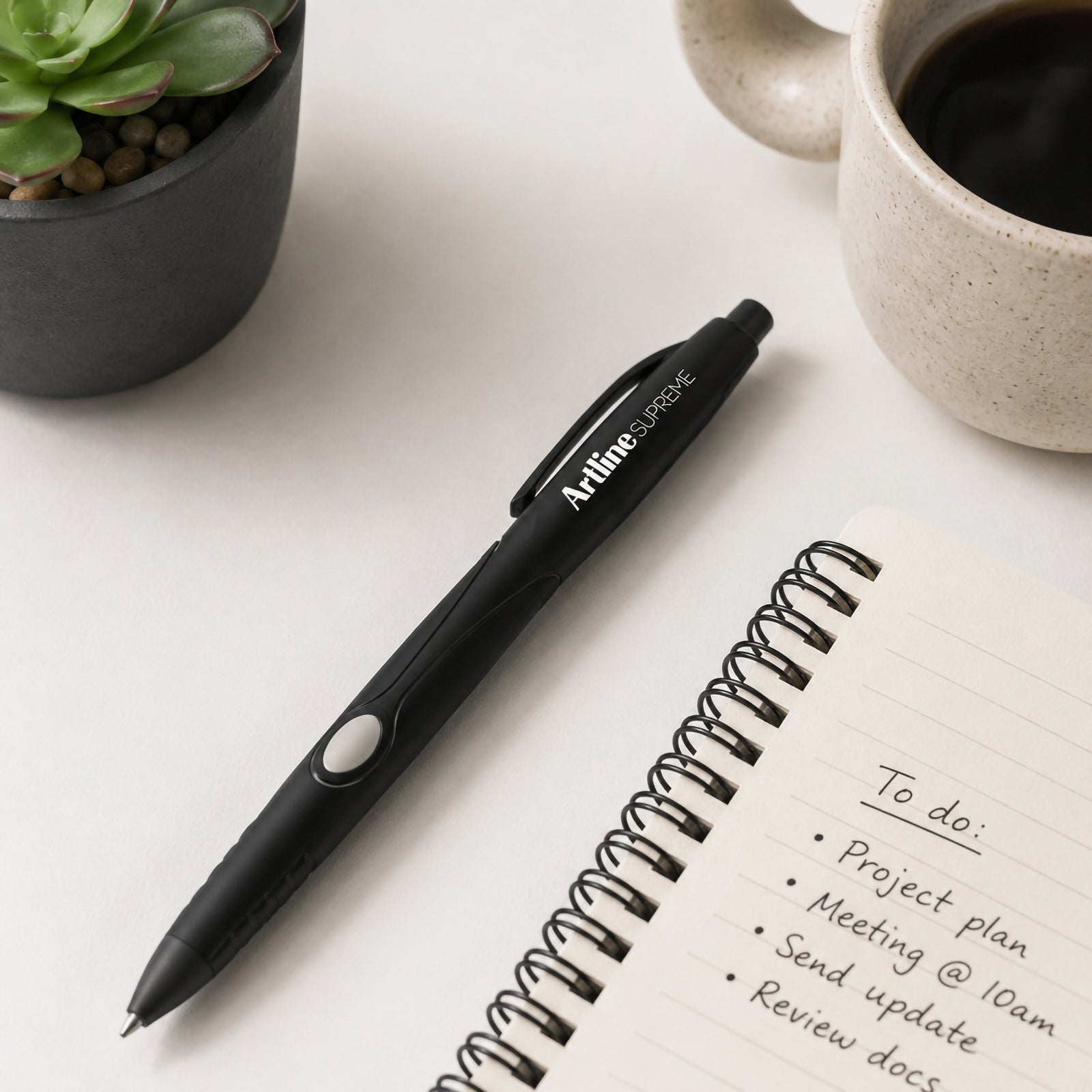 The Artline Supreme Retractable Ballpoint Pen 1.0mm Black is displayed on a clean white desktop beside a spiral notebook, coffee mug and decorative succulent. The image showcases the premium matte black barrel, retractable push button, contoured comfort grip and stylish professional design suitable for everyday office use.