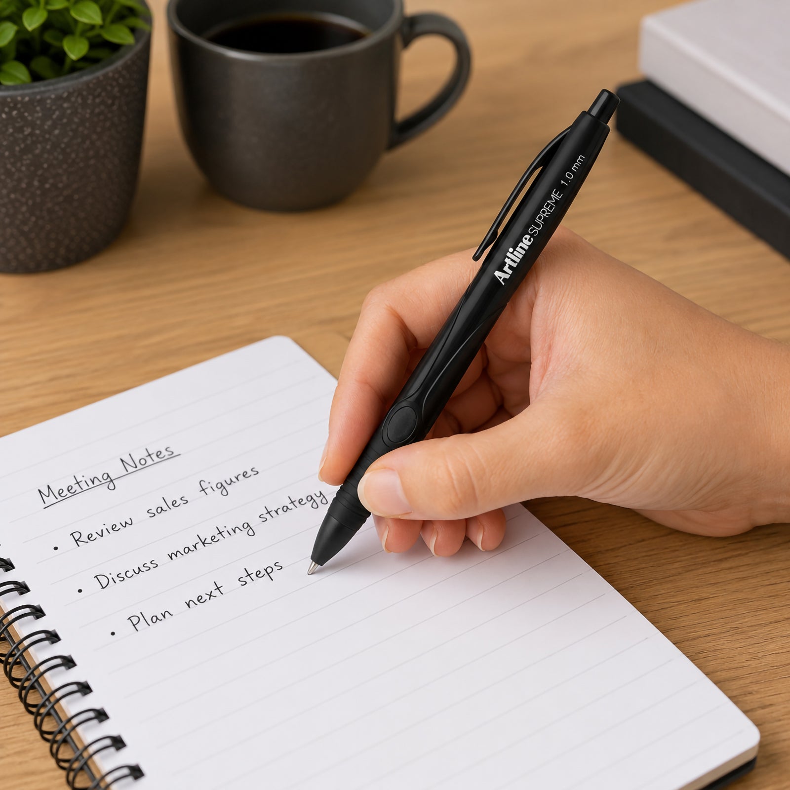 A close up lifestyle image of a person writing meeting notes in a spiral notebook using the Artline Supreme Retractable Ballpoint Pen 1.0mm Black. The ergonomic soft grip and retractable design are clearly visible on a modern wooden desk with a coffee mug and indoor plants, highlighting comfortable everyday writing for office meetings and note taking.