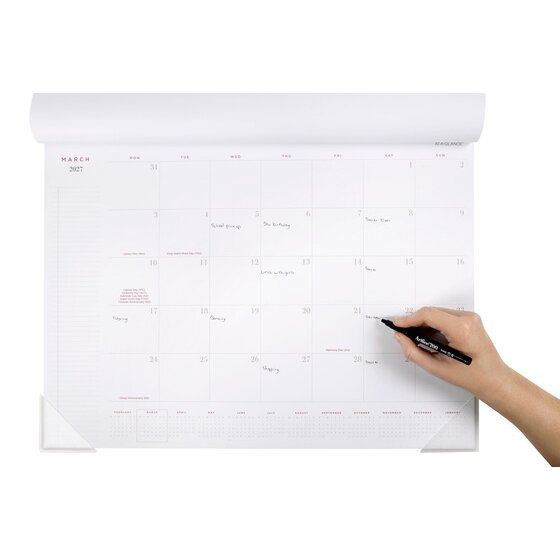 At-A-Glance AAG100127 desk pad open at March 2027 with handwritten entries and a hand writing with a black marker, Monday to Sunday columns and clear corner protectors.