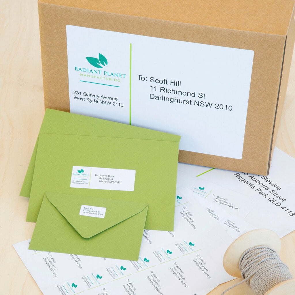 Avery address labels in use, a white printed address label applied to the front of a brown kraft shipping carton, two green envelopes each carrying a smaller printed address label, and part-used label sheets and a spool of twine on a pale timber bench