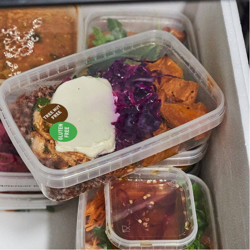 Clear takeaway containers of prepared meals stacked in a commercial fridge, with round Avery allergy labels applied to the lids, a brown Tree Nut Free and a green Gluten Free sticker clearly visible on the front container