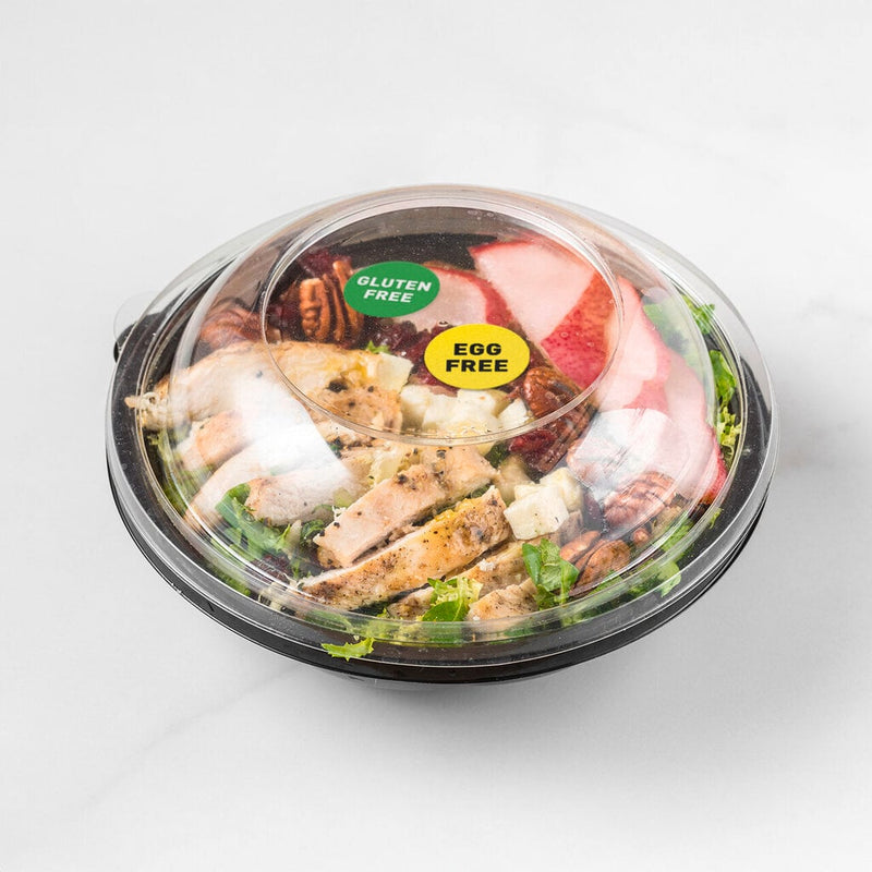 Black round takeaway salad bowl with a clear domed lid on a white bench, labelled with a green Gluten Free and a yellow Egg Free round Avery allergy sticker applied side by side on the lid