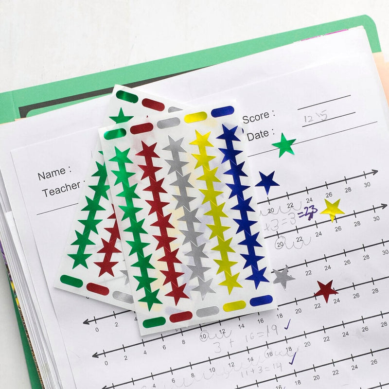 Two sheets of foil star stickers in green, red, silver, yellow and blue resting on a marked maths worksheet in a green folder, with several stars already stuck beside the completed sums