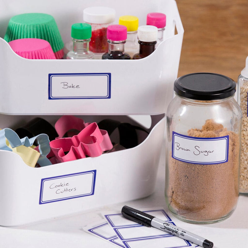 White labels with a blue printed border in use on kitchen storage, two white tubs marked Bake and Cookie Cutters in handwriting, a glass jar marked Brown Sugar and a black marker alongside