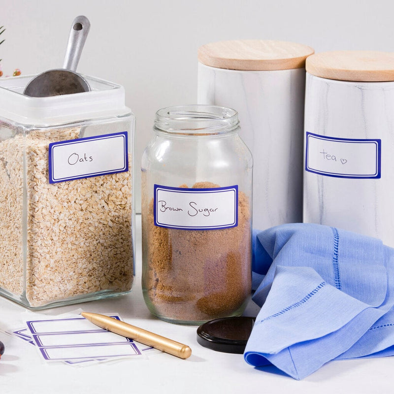 A pantry shelf of glass and marble canisters labelled Oats, Brown Sugar and Tea on blue-bordered white stickers written by hand, with a gold pen and spare blank labels in the foreground