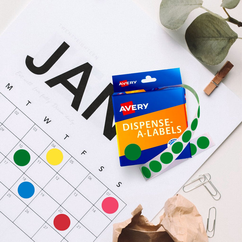 Flat lay of a January wall calendar with dates marked by green, yellow, blue, pink and red round stickers, beside an Avery Dispense-A-Labels carton dispensing a roll of green dots, paper clips and a leafy sprig.