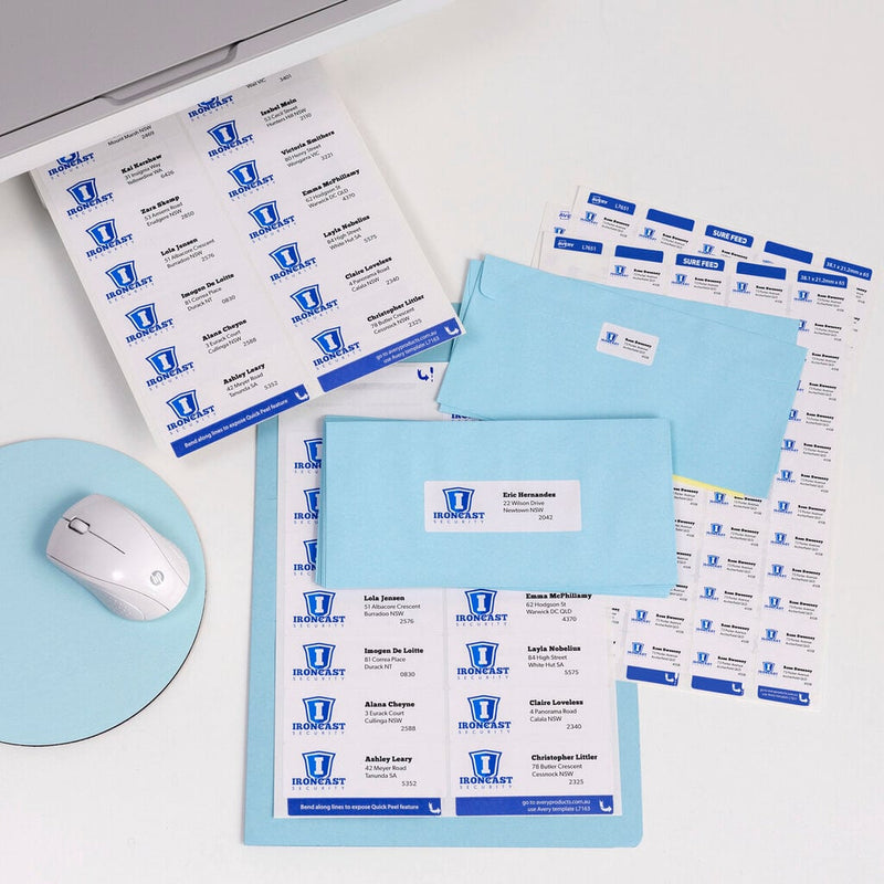 Desk with sheets of printed Avery address labels carrying a blue Ironcast logo, light blue envelopes with labels applied, a mouse and a printer edge.