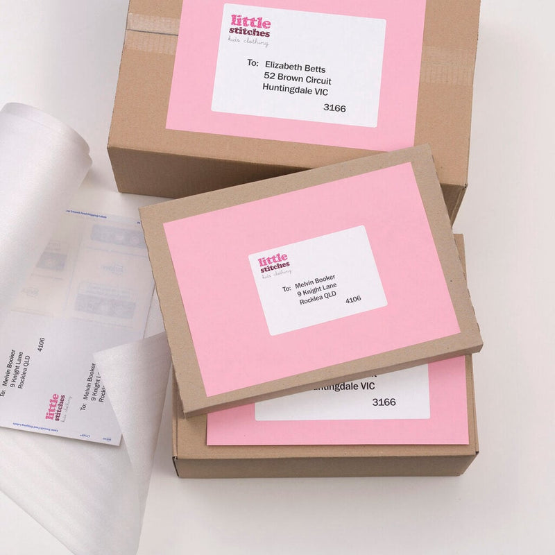 Brown cartons with pink paper panels and white Avery shipping labels applied, beside a printed label sheet on a white desk.