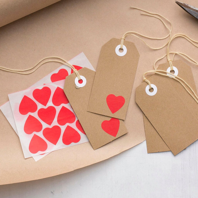Red heart stickers on their backing sheet spread over kraft brown paper next to luggage-style gift tags strung with cord, hearts applied to the tags as a simple decorative finish