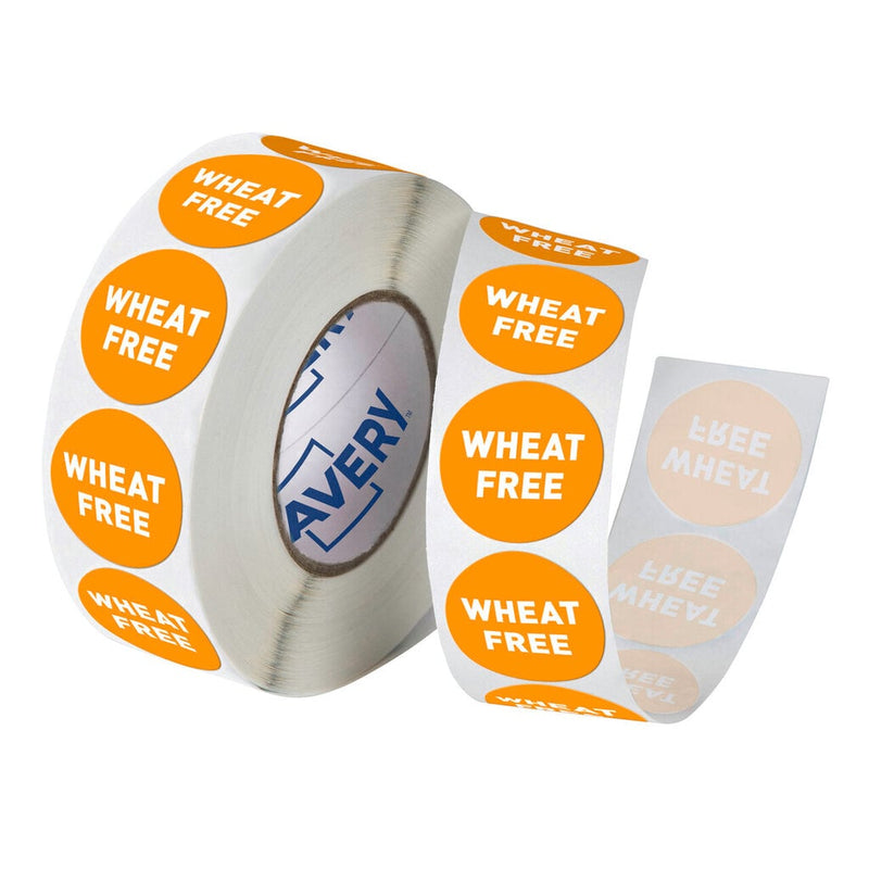 Roll of Avery 937396 Wheat Free allergy labels, orange round 24mm stickers with bold white WHEAT FREE text wound on a white backing roll with a blue Avery logo visible on the core, and a short peeled strip showing the labels lifting from the liner