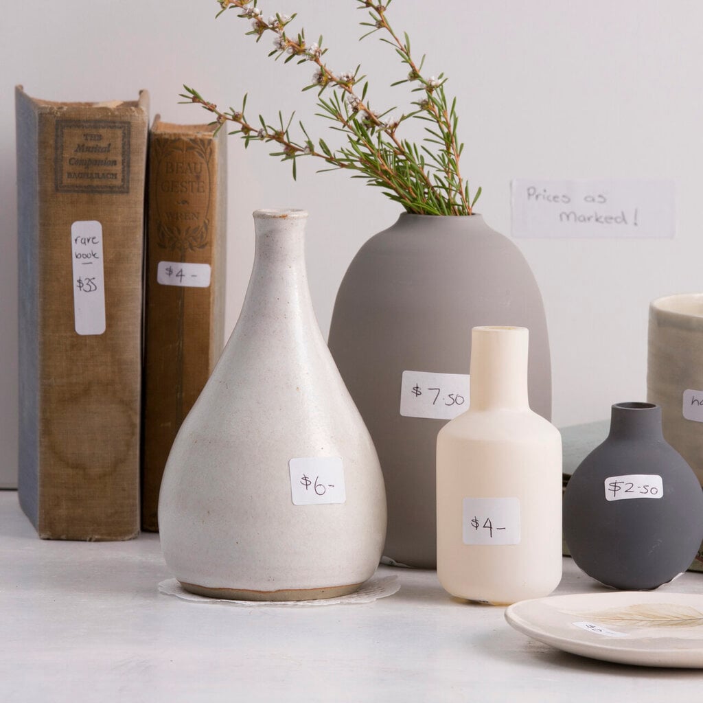 Range application picture of homewares and old books arranged on a light bench, individually priced with small handwritten white stickers, with a native flowering stem in a grey vase behind.