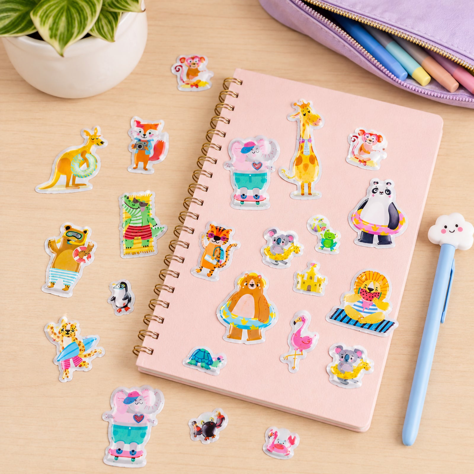 Pink notebook decorated with colourful Beach Buddies puffy stickers featuring cheerful animal characters enjoying beach activities. Designs include giraffes, koalas, kangaroos, flamingos, pandas and lions with float rings, surfboards and summer accessories.