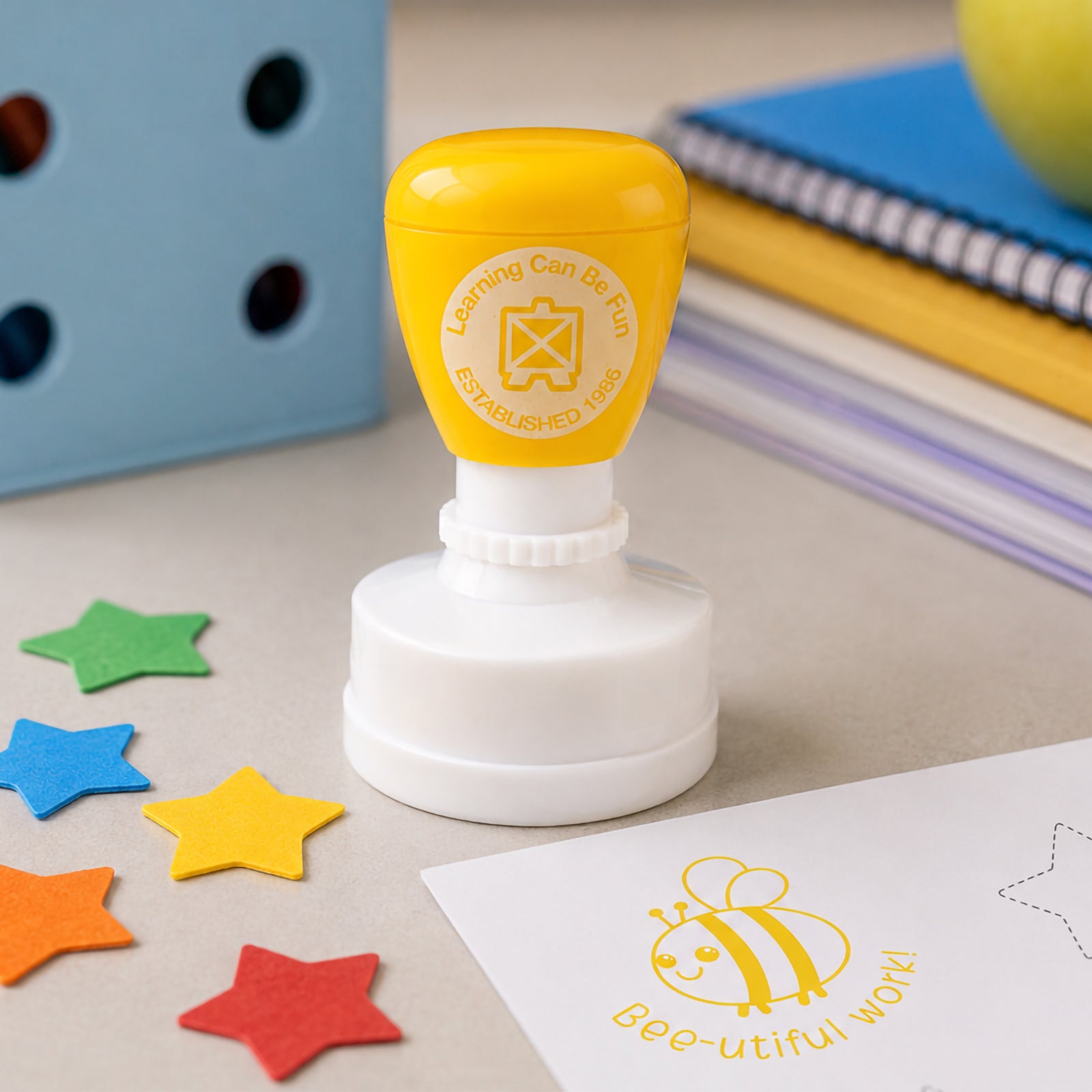 Close up classroom scene featuring the self inking merit stamp with colourful star shapes, stationery and educational materials. The bright yellow impression helps reward effort, improvement and participation in a fun and memorable way.