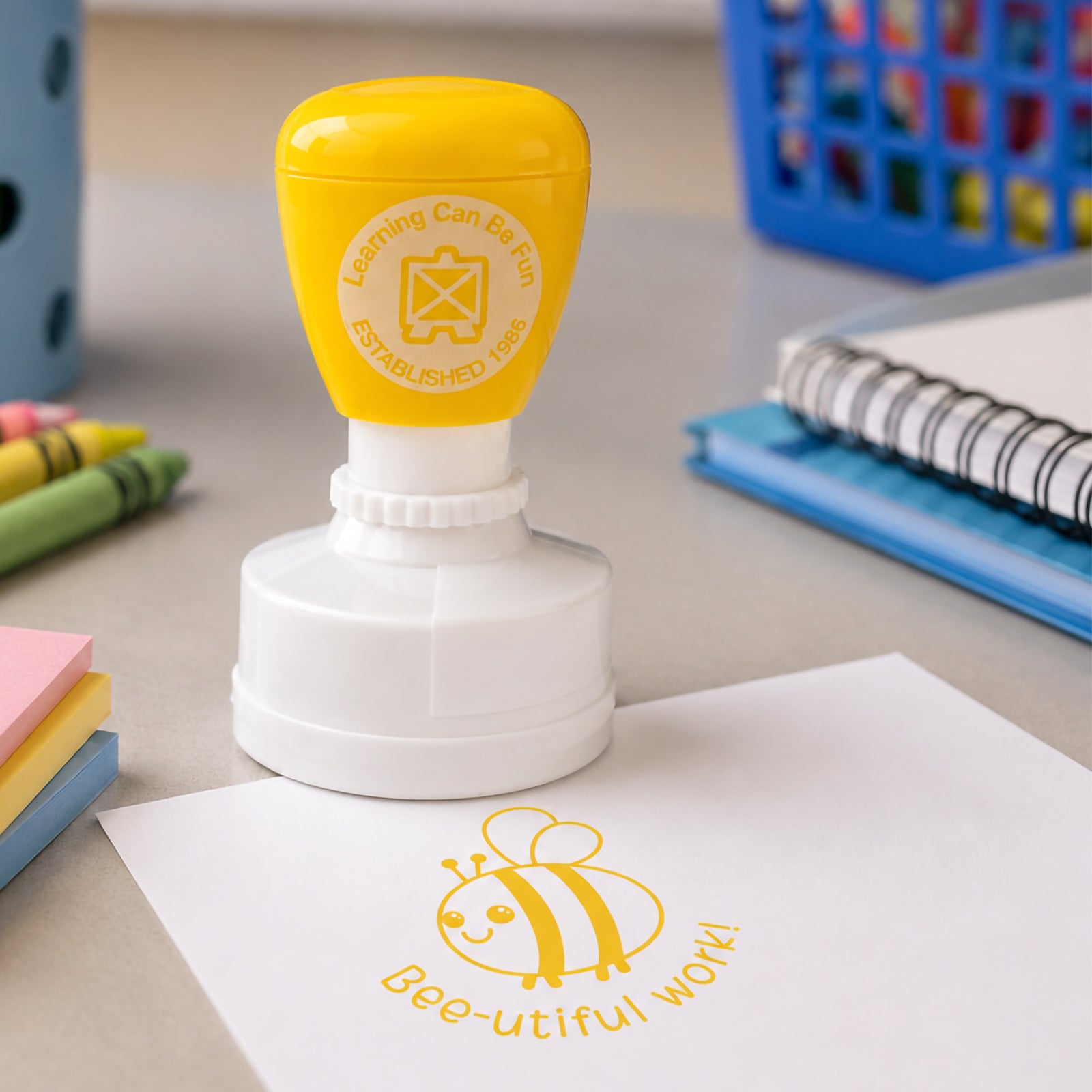 Yellow Bee-utiful Work reward stamp displayed beside sticky notes, crayons and exercise books. The cheerful bee design and motivational message demonstrate an engaging way to provide classroom feedback and celebrate student achievement.